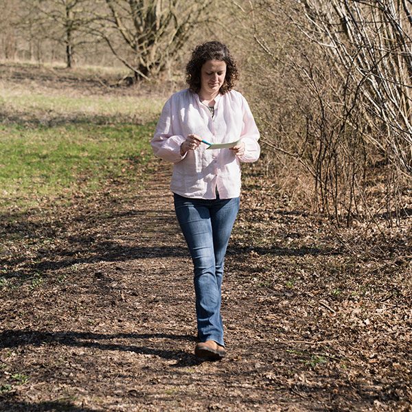 Walk and Talk - Therapie im Gehen Systemische Beraterin mit Karte, die einen Waldweg entlanggeht, umgeben von Bäumen und grüner Wiese.