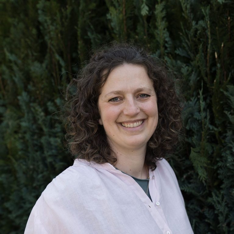 Systemische Beraterin Flensburg Freundliche Frau mit lockigem Haar vor grünen Pflanzen.